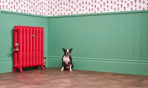 Ornate Rococo II cast iron radiator in Heritage Red on feet, with a valve, against green panelling with a dog on a parquet floor.