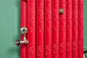 Close-up of an ornate Rococo II cast iron radiator in Benjamin Moore Heritage Red HC181 with a brass thermostatic valve against a green panelled wall.