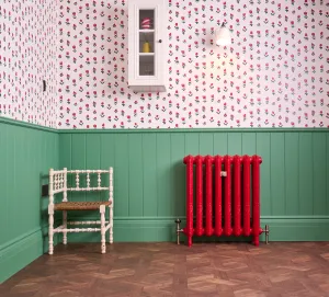 Ornate Rococo II cast iron radiator in red on feet, with brass valves, against green panelling beside a white corner chair on parquet.