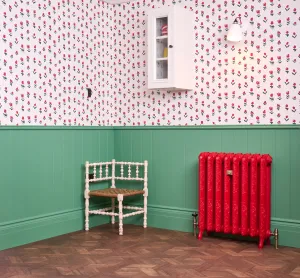 Ornate Rococo II cast iron radiator in Heritage Red HC181 on feet with brass valves, against green panelling beside a white corner chair.