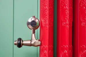 Close-up of ornate Rococo II cast iron radiator columns in Benjamin Moore Heritage Red HC181 with a chrome air valve on a brass-finish fitting, green panelled wall.