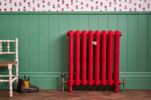 Ornate Rococo II cast iron radiator in Heritage Red on feet, with brass valves, against green panelling beside a white chair and boots.