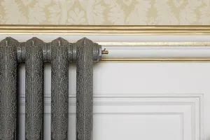 Close-up of an ornate Rococo II cast iron radiator in a grey crackle finish with a brass bleed valve, against white panelling and gilded moulding.