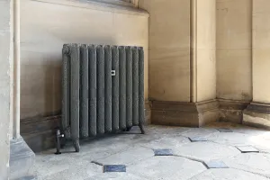 Ornate Rococo II cast iron radiator in a dark grey crackle finish on feet, with a black valve, set against a sandstone wall on flagstones.