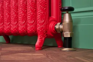 Close-up of an ornate Rococo II cast iron radiator in Benjamin Moore Heritage Red HC181 on feet, with a brass Windsor valve, green wall and parquet floor.