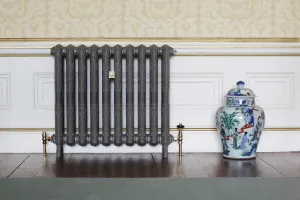 Ornate Rococo II 760mm cast iron radiator on feet in a dark metallic grey finish with brass valves, beside a porcelain jar on a wooden floor.