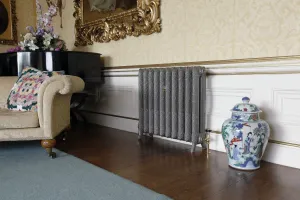 Ornate Rococo II 760mm cast iron radiator in a pewter grey finish on feet with a brass valve, by a sofa, piano and porcelain jar.