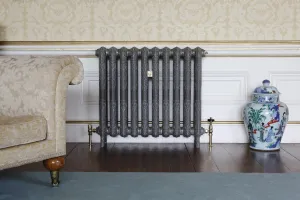 Ornate Rococo II cast iron radiator in burnished dark grey with brass valves, between a damask sofa and a blue-and-white jar on dark floorboards.