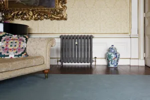Ornate Rococo II cast iron radiator in dark metallic grey on feet with brass valves, below damask wallpaper beside a sofa and ginger jar.