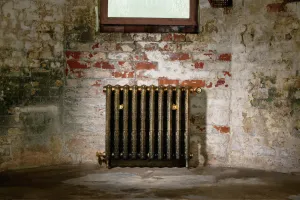 Ornate Rococo II cast iron radiator in Aged Gold on feet, with a Windsor angled valve, against a whitewashed brick wall below a window.