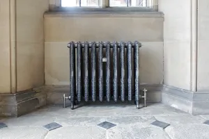 Ornate Rococo II cast iron radiator in Full Polish on feet, with angled valves, beneath a window against a stone wall on a flagstone floor.