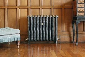 Ornate Rococo II cast iron radiator on feet, dark with polished raised pattern, brass valves, against oak panelling by a footstool and cabinet.