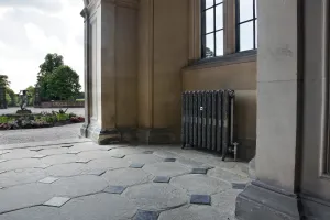 Ornate Rococo II cast iron radiator in Full Polish on feet, below a window in a stone loggia with flagstone floor and garden beyond.