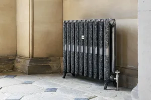 Ornate Rococo II cast iron radiator in Full Polish on feet, with a thermostatic valve, against a sandstone wall on a flagstone floor.