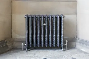 Ornate Rococo II cast iron radiator on feet in Full Polish, with silver valves at each end, in a stone alcove on a flagstone floor.