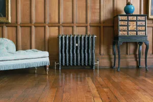 Ornate Rococo II cast iron radiator on feet, dark with polished highlights and nickel valves, against oak panelling on a wooden floor.