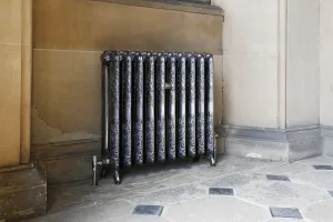 Ornate Rococo II cast iron radiator in Full Polish on feet, with a valve, against a sandstone wall on a flagstone floor.
