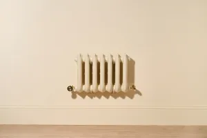 Ornate seven-column Rococo III cast iron radiator in Farrow & Ball Matchstick No. 2013, wall-hung with Natural Brass valves above a skirting board.