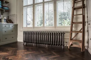 Ornate Rococo III cast iron radiator in Full Polish on feet beneath a shuttered window, with a library ladder on a herringbone floor.