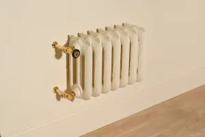 Ornate wall-hung Rococo III cast iron radiator in Farrow & Ball Matchstick No. 2013 with Natural Brass valves, above a skirting board and oak floor.