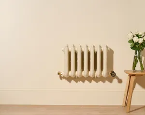 Ornate seven-column Rococo III radiator in Farrow & Ball Matchstick, wall-hung with Natural Brass valves, beside a stool with white roses.