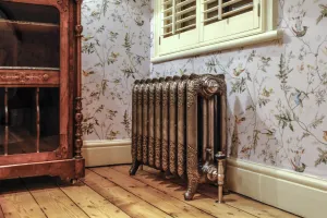 Ornate Rococo III cast iron radiator on feet in Full Polish with a nickel valve, below a shuttered window against bird-print wallpaper.