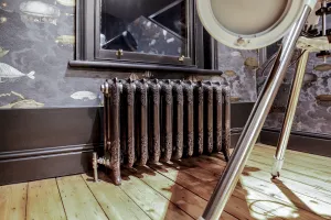 Ornate Rococo III cast iron radiator in Full Polish on feet below a sash window, with angled valve, dark skirting and pine floorboards.