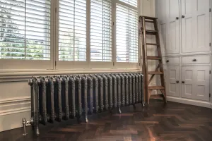 Ornate Rococo III cast iron radiator in Full Polish on feet below shuttered windows, with an angled valve, herringbone floor and wooden ladder.