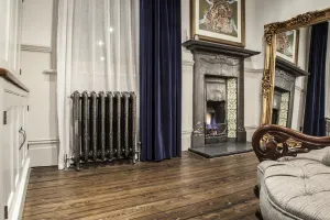 Ornate Rococo III cast iron radiator in Full Polish on feet below a curtained window, beside a cast iron fireplace on wooden floorboards.