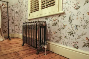 Ornate Rococo III cast iron radiator in Full Polish on feet with a polished valve, below a shuttered window on bird-print wallpaper and pine floorboards.