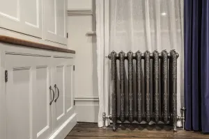 Ornate Rococo III cast iron radiator in Full Polish on feet with angled valves, by a sheer curtain, navy drape and painted cupboards.