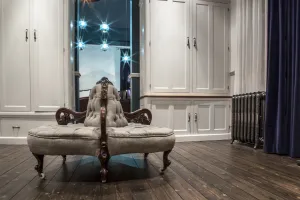 Ornate Rococo III cast iron radiator in Full Polish on feet beside navy curtains, with a tufted grey round settee on dark floorboards.