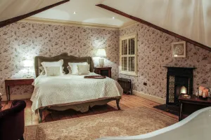 A low Rococo III cast iron radiator in a dark finish under a shuttered window in an attic bedroom with floral wallpaper and a fireplace.