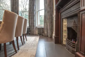 Two ornate Rococo III cast iron radiators in Full Polish on feet beneath a curtained bay window, beside dining chairs and a tiled cast iron fireplace.