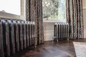 Two ornate Rococo III cast iron radiators in Full Polish on feet beneath a bay window, with patterned curtains and a herringbone wood floor.
