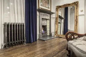 Ornate Rococo III 470mm cast iron radiator in Full Polish on feet by a curtained window, near a tiled fireplace and gilt mirror.