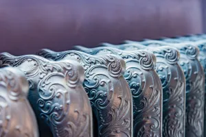 Close-up of the ornate scrolled tops of a Rococo III cast iron radiator in a polished silver metallic finish against a purple wall.