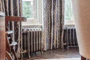 Two ornate Rococo III 470mm cast iron radiators in Full Polish on feet beneath bay windows, with patterned curtains and herringbone floor.