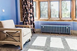Ornate Rococo III 470mm cast iron radiator in a pale blue metallic finish on feet beneath a stone mullioned bay window, beside an armchair.
