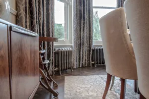 Two ornate Rococo III cast iron radiators in Full Polish on feet beneath bay windows with patterned curtains, herringbone floor and dining chairs.