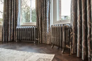 Two ornate Rococo III cast iron radiators in Full Polish on feet beneath a bay window, between patterned curtains on a herringbone floor.