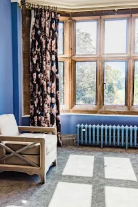 Low Rococo III cast iron radiator in pale blue on feet beneath a stone mullioned bay window, with floral curtains and an armchair.