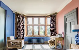 Low Rococo III cast iron radiator in pale blue-grey on feet with valves, beneath a mullioned bay window in a blue and pink bedroom.