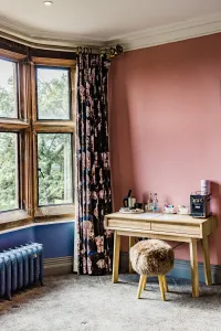 Ornate Rococo III 470mm cast iron radiator in blue on feet beneath a bay window, beside floral curtains, a pink wall and a wooden desk.
