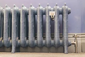 Ornate Rococo III 470mm cast iron radiator in a blue-grey finish on feet, with a nickel angled valve and wall stay, against a lilac wall.