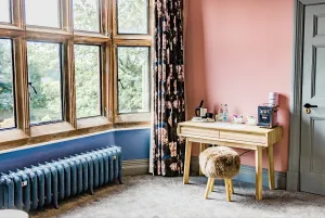Ornate Rococo III 470mm cast iron radiator in blue-grey on feet beneath a stone mullioned window, beside a desk in a pink-walled bedroom.