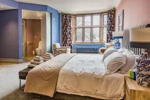 A low Rococo III cast iron radiator in a pale blue finish beneath a mullioned bay window in a bedroom with a double bed and armchair.