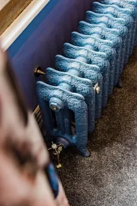 Ornate Rococo III 470mm cast iron radiator in a mid-blue finish on feet, with brass valves, against a blue wall on grey carpet.