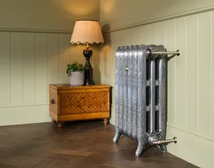 Ornate Rococo III cast iron radiator in Full Polish on scrolled feet with brass-coloured pipes, beside a parquetry chest and lamp on herringbone floor.