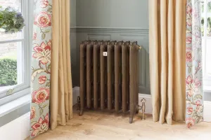 Ornate Rococo III cast iron radiator in Beaten Brass on feet with angled valves, set in a window bay between floral curtains on an oak floor.